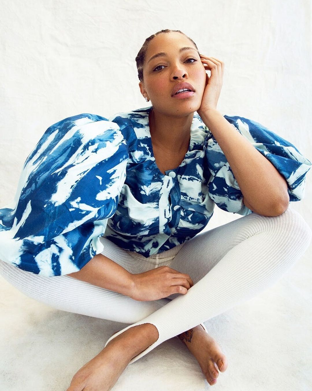 Portrait of an adult woman sitting cross-legged and posing. She is wearing a blue and white blouse with white leggings and is barefoot.
