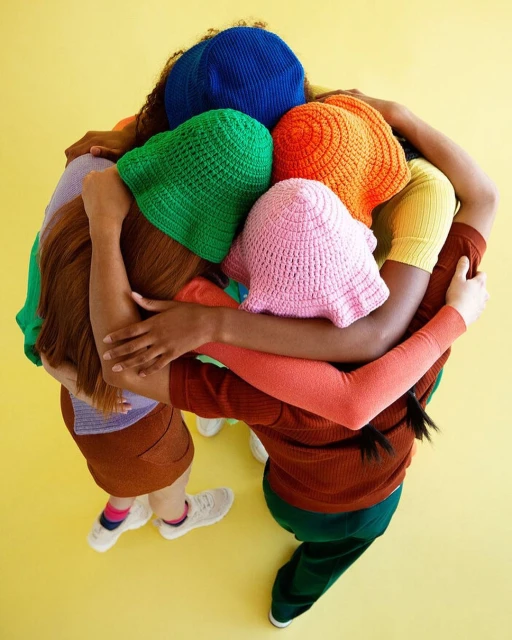 Overhead shot of a group of diverse adults in a circle hugging, wearing colorful crocheted hats against a bright yellow background.