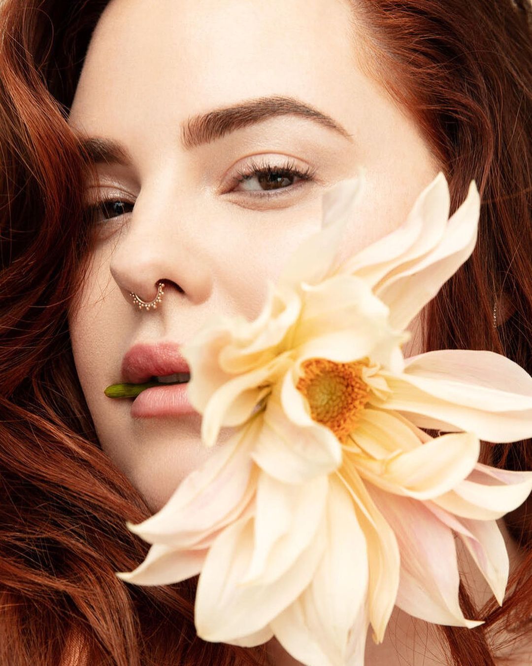 Close-up portrait of a red-haired woman with a nose ring, holding a large cream-colored flower in her mouth, in a beauty context.