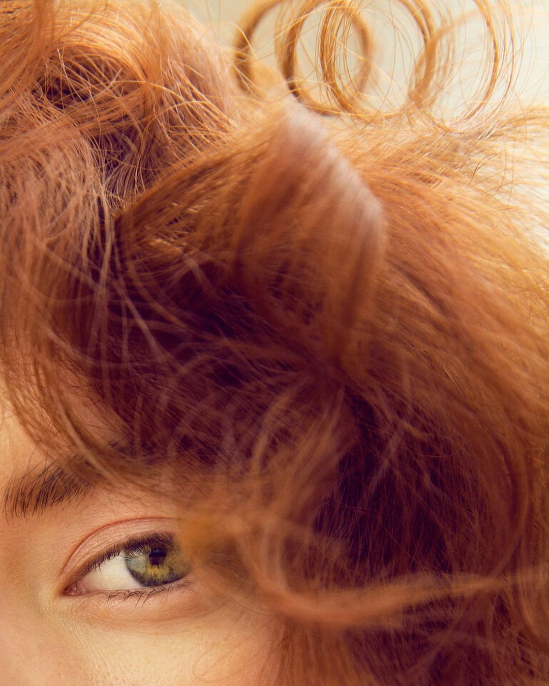 Close-up portrait of an adult woman with auburn curly hair partially obscuring her face, emphasizing her green eye against a soft backdrop.