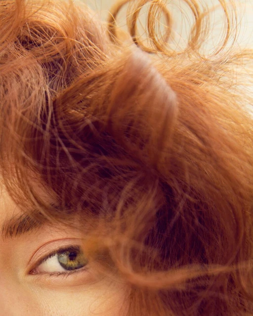 Close-up portrait of an adult woman with auburn curly hair partially obscuring her face, emphasizing her green eye against a soft backdrop.