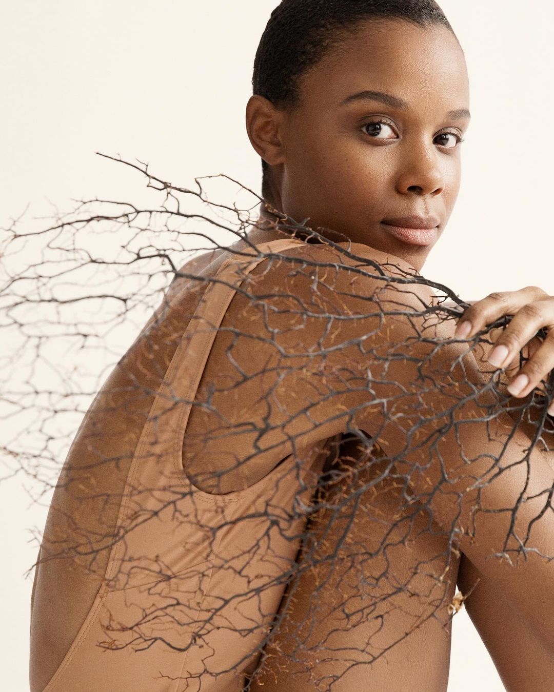 Close-up studio portrait of a Black woman looking over her shoulder, holding bare tree branches against a neutral background.