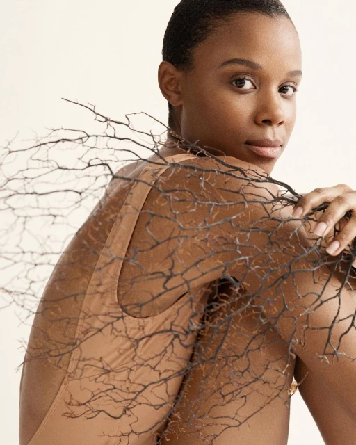 Close-up studio portrait of a Black woman looking over her shoulder, holding bare tree branches against a neutral background.