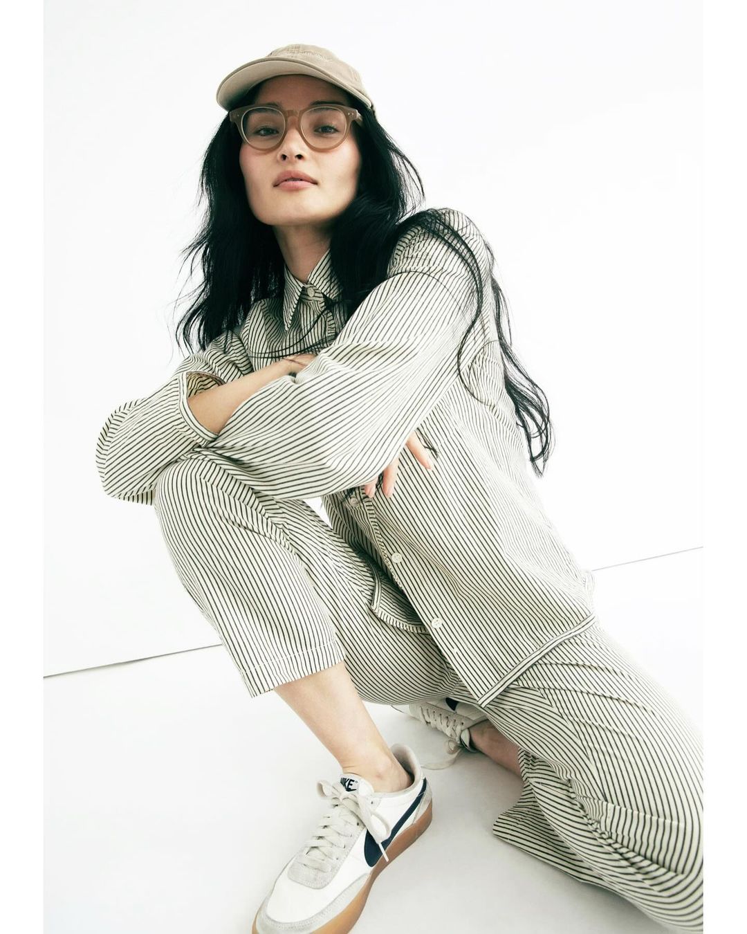 Woman in striped shirt and pants crouches indoors with white Nike sneakers and a beige baseball cap, shot at eye-level.