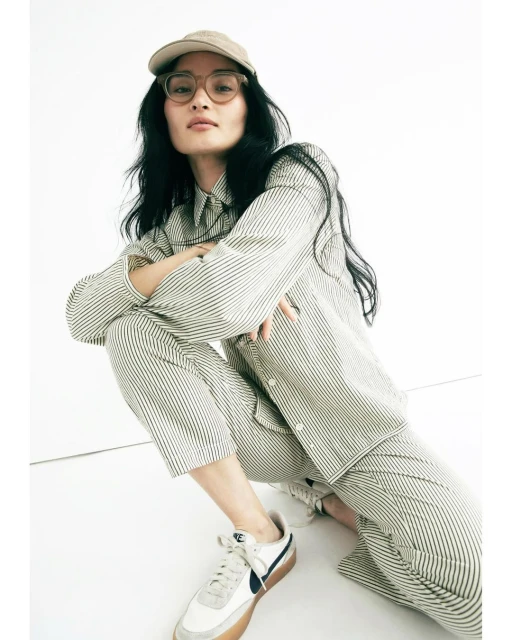 Woman in striped shirt and pants crouches indoors with white Nike sneakers and a beige baseball cap, shot at eye-level.