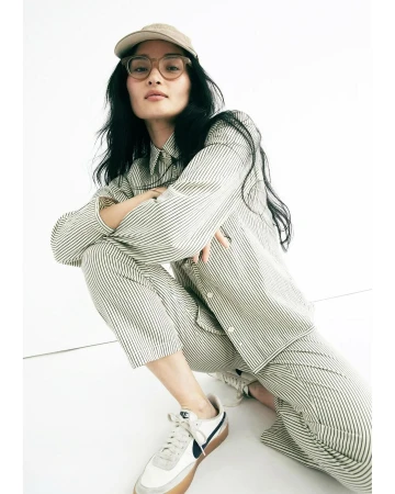 Woman in striped shirt and pants crouches indoors with white Nike sneakers and a beige baseball cap, shot at eye-level.