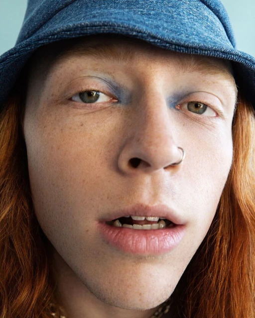 Close-up portrait of a person with red hair, wearing a denim hat and light blue eyeshadow, facing forward in a well-lit indoor studio.