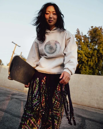East Asian woman holding a skateboard wearing a cream hoodie and patterned pants outdoors on an asphalt street at sunset