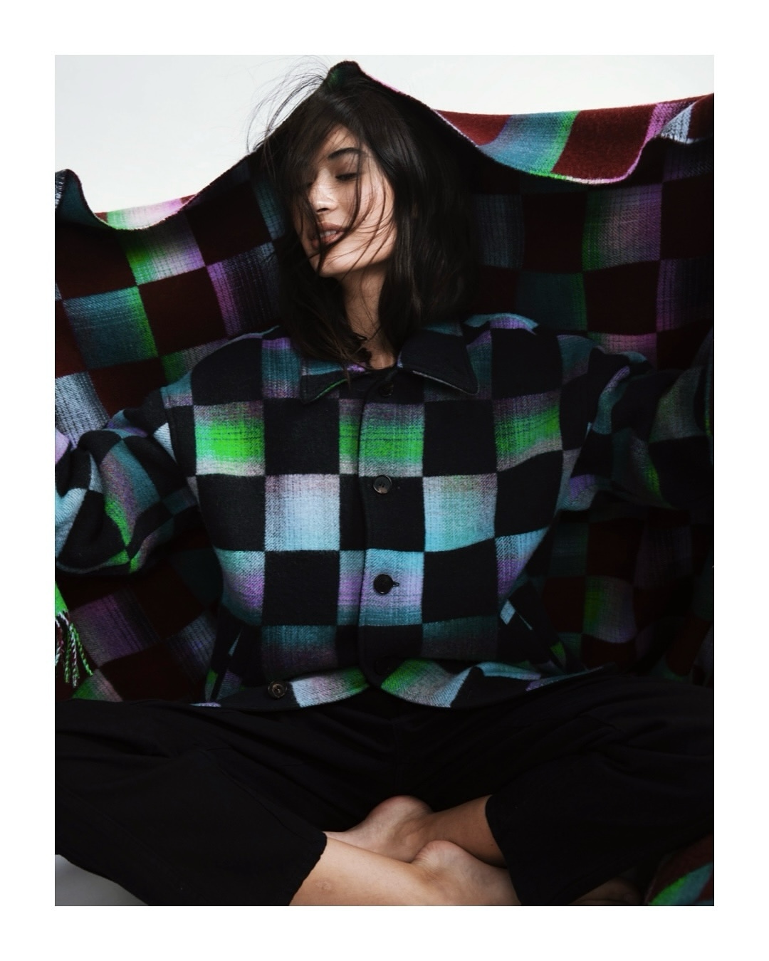 A woman with dark hair sits with crossed legs, wearing a multicolored checkered jacket and black pants against a neutral background.