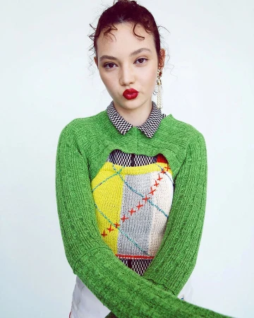 Portrait of a woman wearing a green textured cropped sweater, checkered collar, and red lipstick against a plain white background.