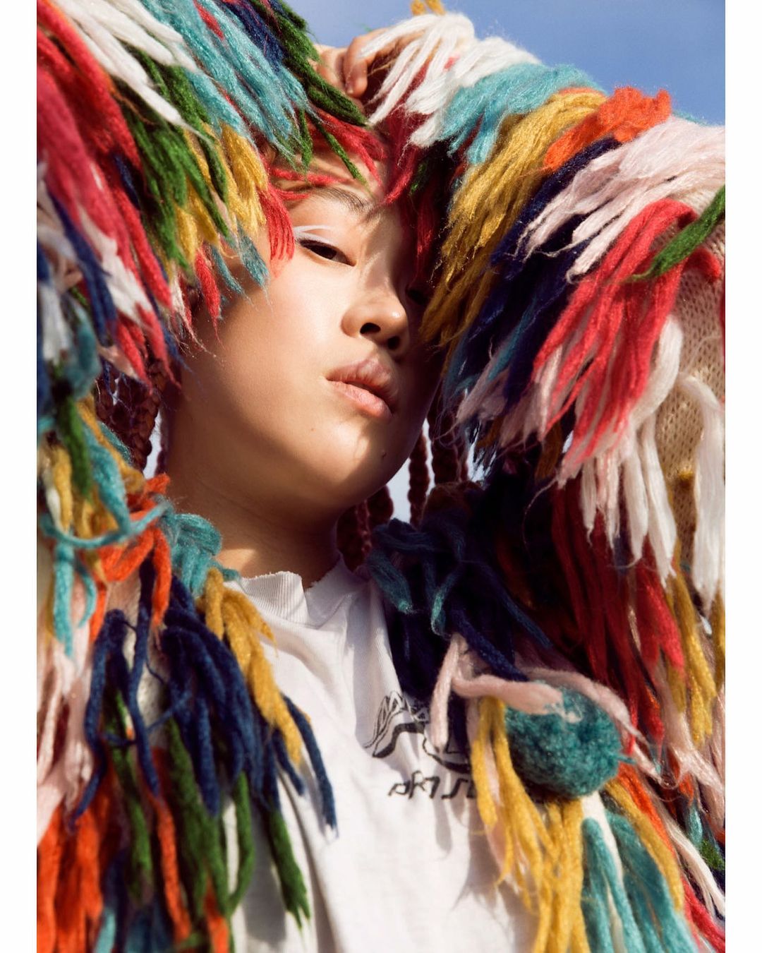 Close-up of a young Asian woman with white eyeliner and a colorful fringed garment, looking serene against a light blue sky.
