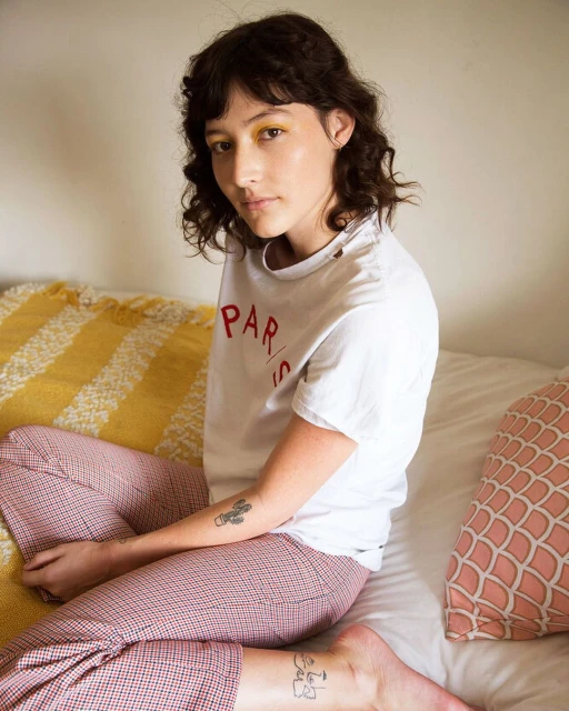 Young woman sitting on a bed with a yellow striped blanket wearing a t-shirt that says 'Paris'. Portrait orientation, medium shot.