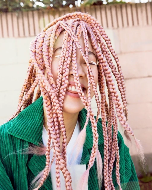 Smiling woman with pink box braids and a green corduroy jacket, eyes closed, standing outdoors with a cheerful expression.