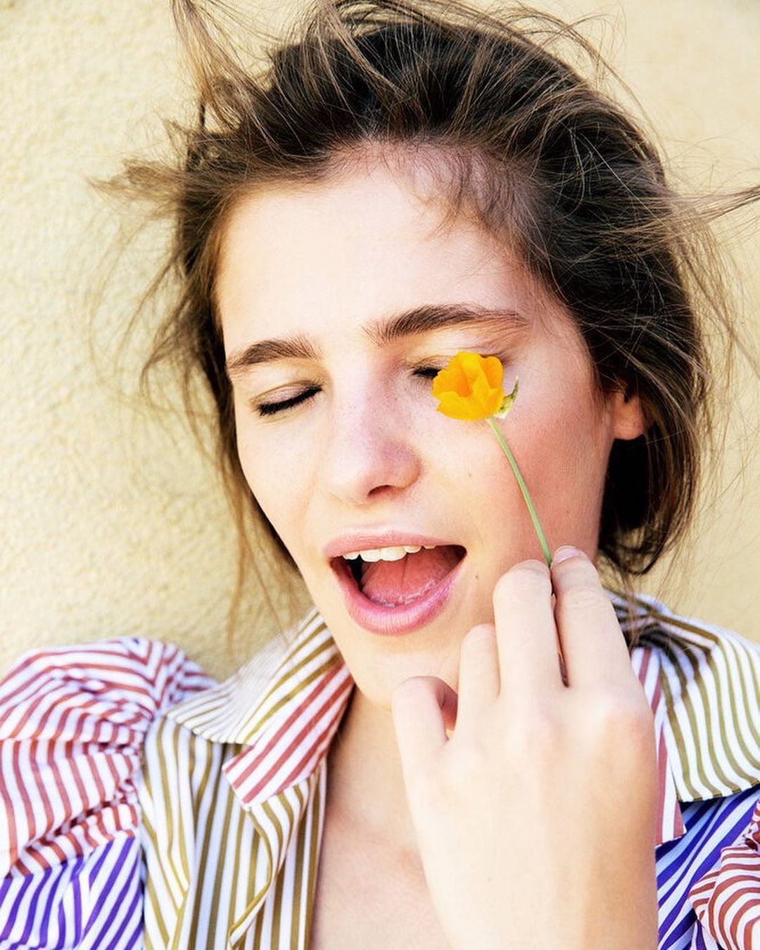 Woman with closed eyes smiles, holding a yellow flower near her face, wearing a striped shirt against a light background in a close-up shot.
