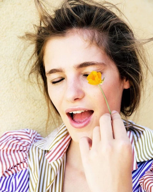 Woman with closed eyes smiles, holding a yellow flower near her face, wearing a striped shirt against a light background in a close-up shot.