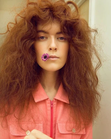 Portrait of a woman with large frizzy reddish-brown hair holding a purple flower in her mouth by photographer Brigitte Sire