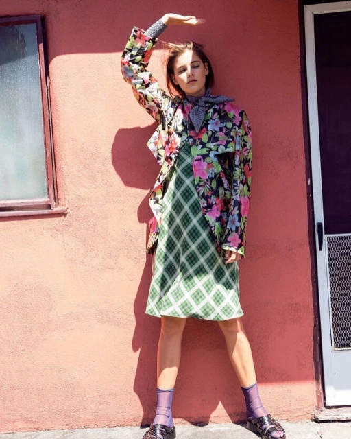 Woman in floral jacket and plaid dress poses against a pink wall with a doorway, showcasing a fashion-forward and retro-inspired style.