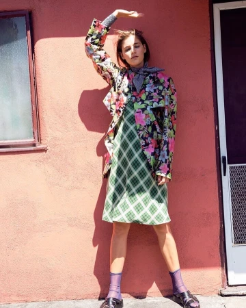 Woman in floral jacket and plaid dress poses against a pink wall with a doorway, showcasing a fashion-forward and retro-inspired style.