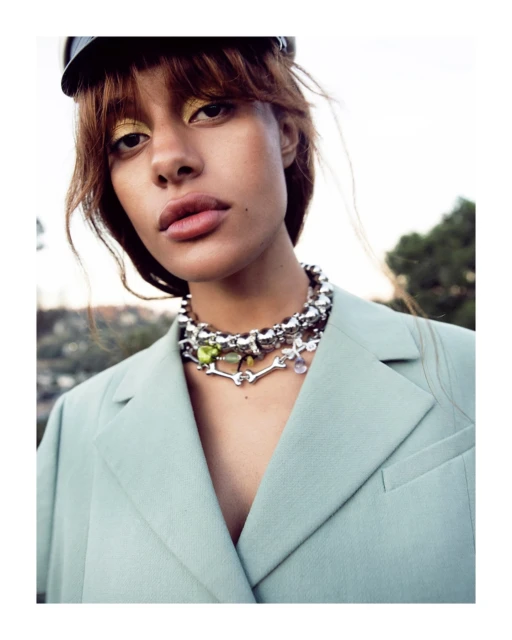 Close-up shot of a stylish adult female wearing a mint-green blazer and a layered silver necklace with colorful gem accents, outdoor setting.
