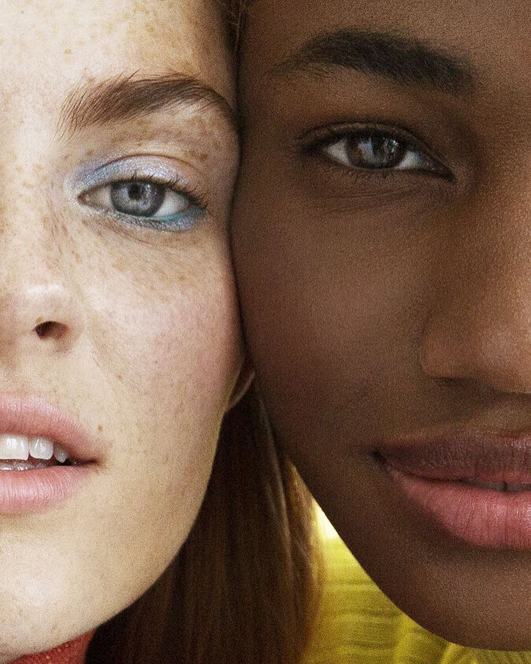 Close-up portrait featuring the faces of two adult women with different skin tones, one fair and freckled, one darker, under studio lighting.
