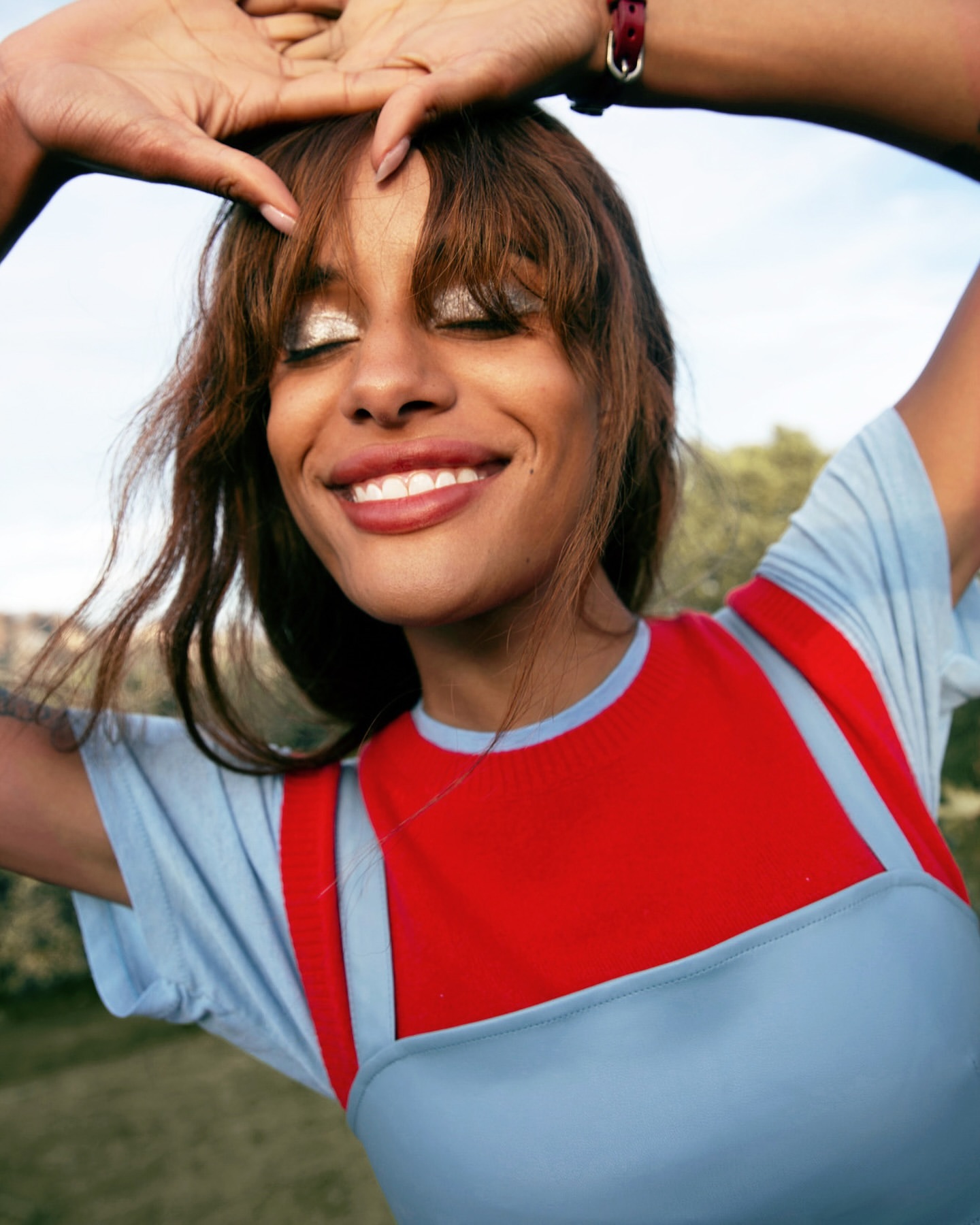 Close-up of a smiling woman with brown hair and light eye makeup, wearing a blue and red top, posing with hands above her head outdoors.