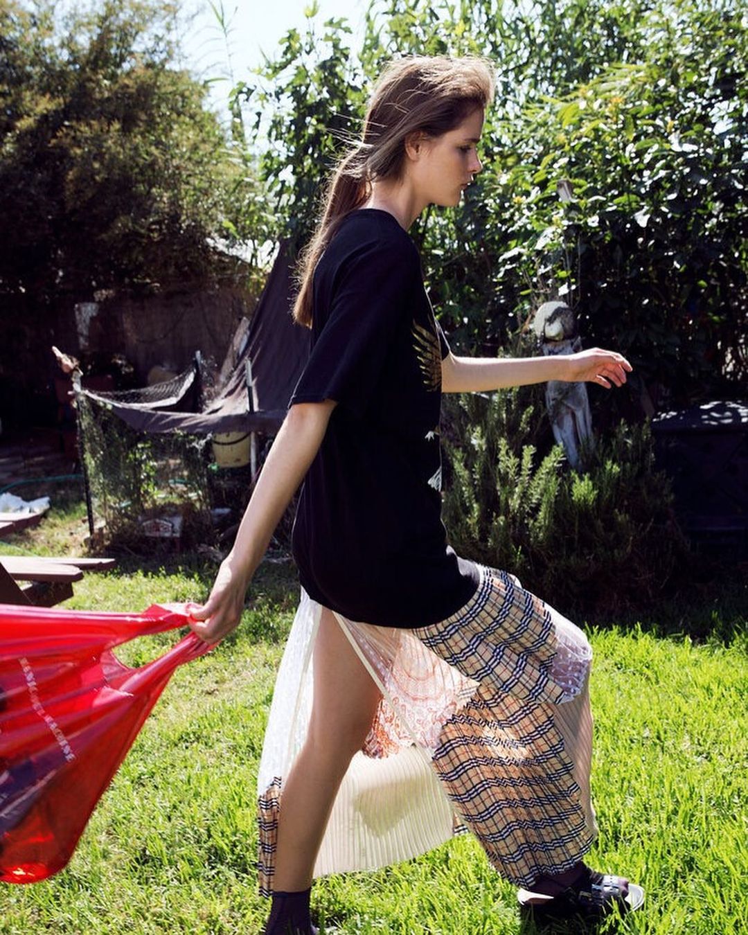 A young adult woman walks outdoors carrying a red shopping bag, wearing a black graphic T-shirt, plaid skirt, and black sneakers.