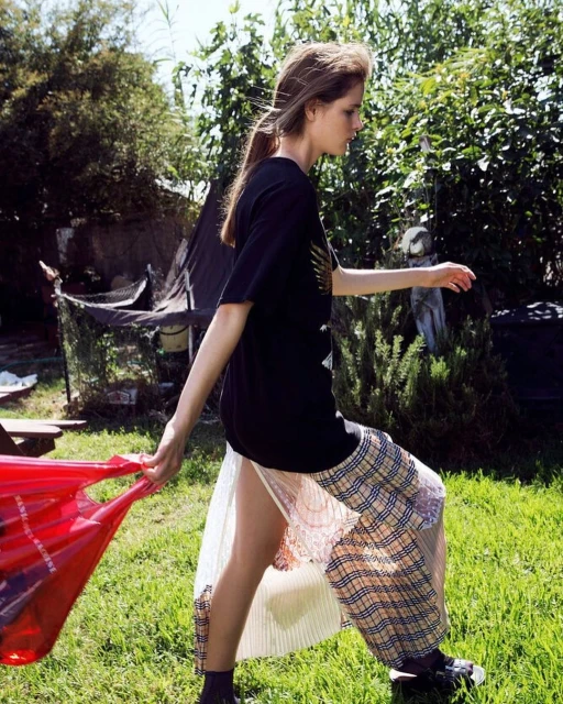 A young adult woman walks outdoors carrying a red shopping bag, wearing a black graphic T-shirt, plaid skirt, and black sneakers.