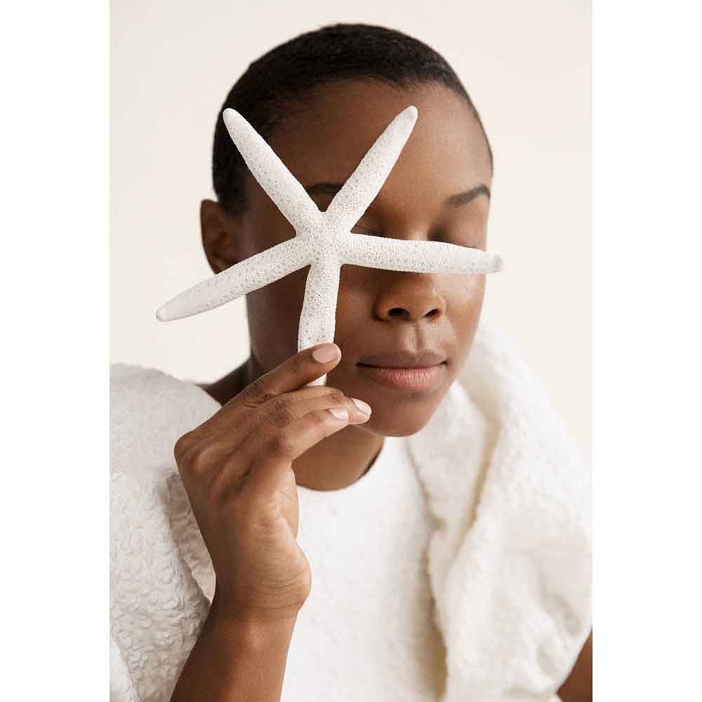 Woman with dark skin poses indoors with white starfish covering her eye, wearing a textured white dress against an off-white background.