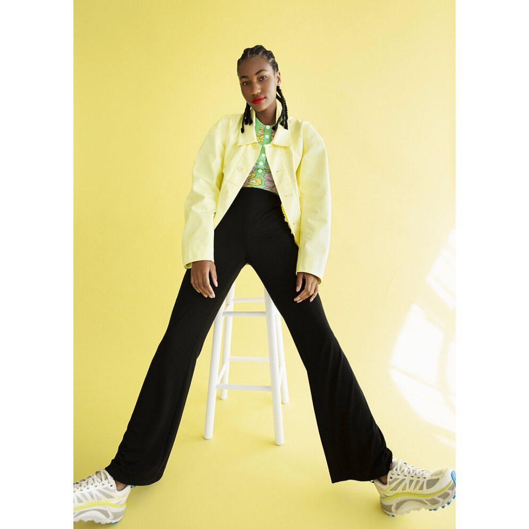 Black woman with braided hair wearing yellow jacket and black pants seated on a white stool against a yellow background