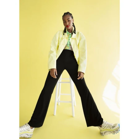 Black woman with braided hair wearing yellow jacket and black pants seated on a white stool against a yellow background