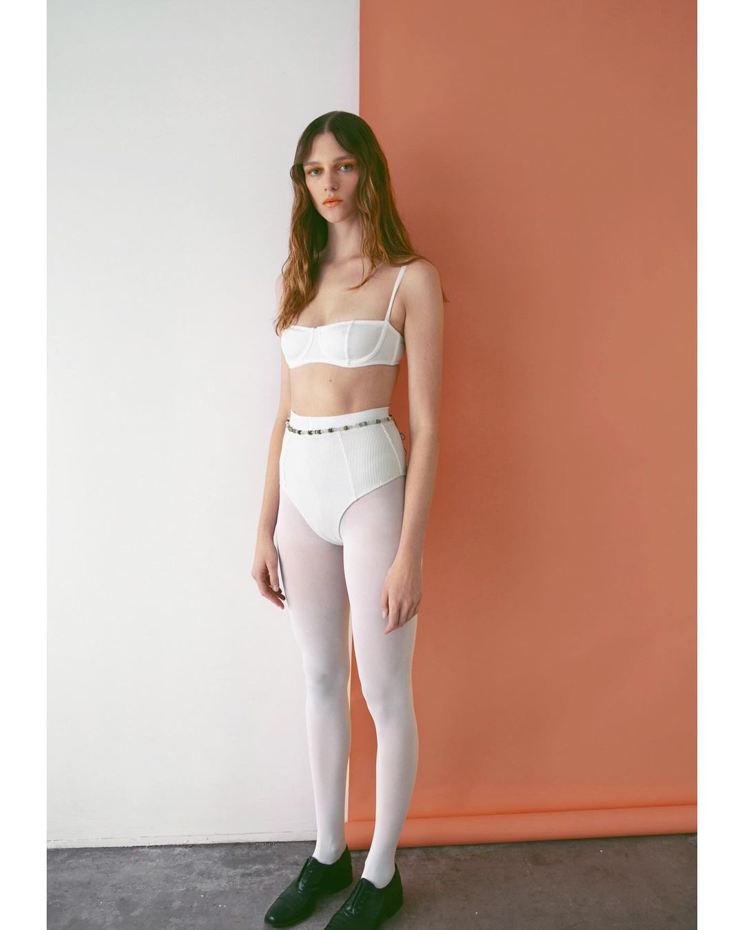 Young woman stands in white lingerie and stockings against a split background of orange and white in a fashion studio setting.