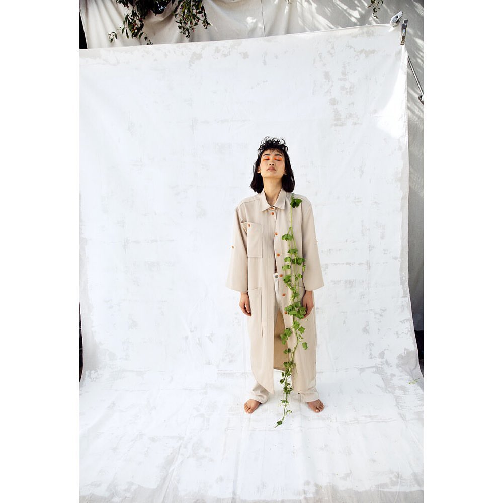 A young adult woman poses in a light-beige jumpsuit with eyes closed and a plant draped on her against a white backdrop in a studio.