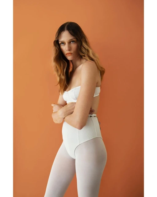Portrait of a fair-skinned female model with wavy brown hair and orange makeup posing in a white bra top and high-waisted briefs.