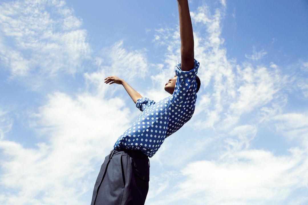 Adult person in a blue polka dot shirt with arms raised against a partly cloudy sky. Low angle shot, fashion editorial.
