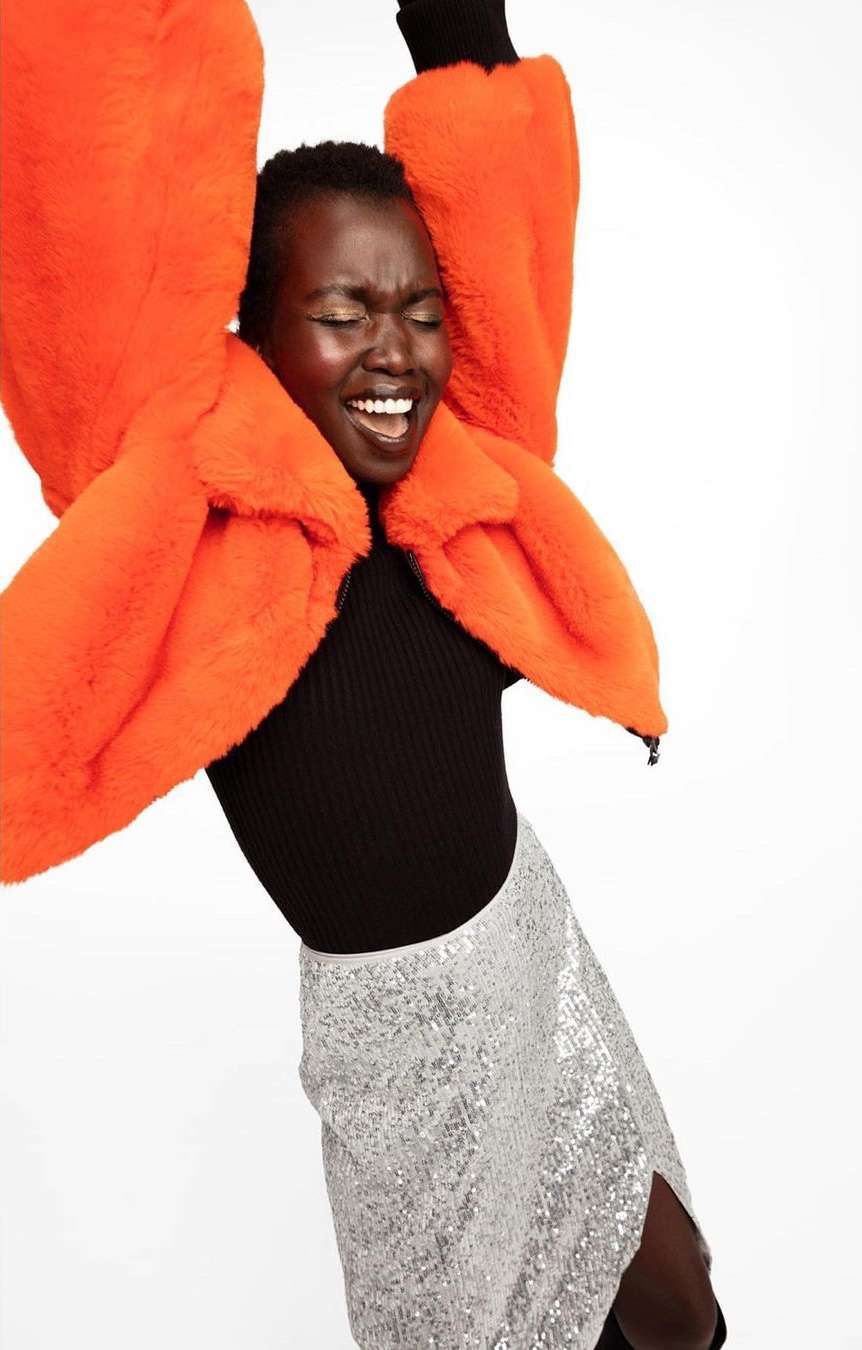 Young woman wearing an orange faux fur coat and sequin skirt smiles with her arms up against a white backdrop in a studio shot.