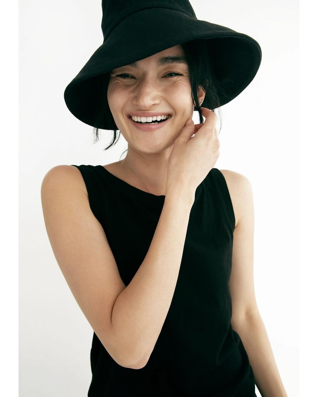 Smiling woman in black tank top and sun hat in a brightly lit indoor space with a white background, head and shoulders shot.