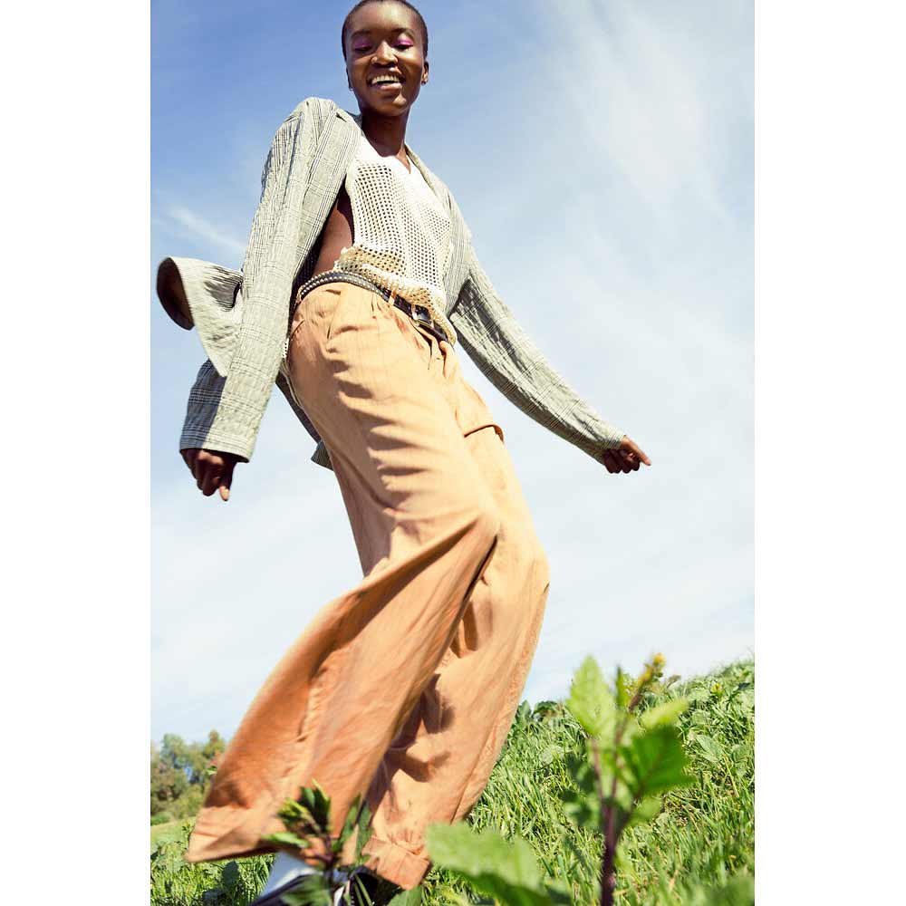 A Black female fashion model with short hair in a long-sleeve shirt and baggy trousers smiles in an outdoor shot on a sunny day.
