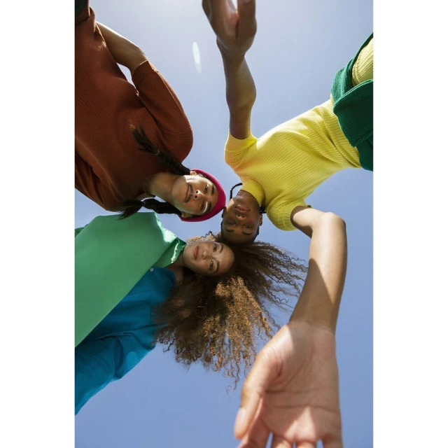 Three young adult women of diverse ethnicities pose together looking down at the camera against a clear blue sky, wearing casual, colorful clothing.