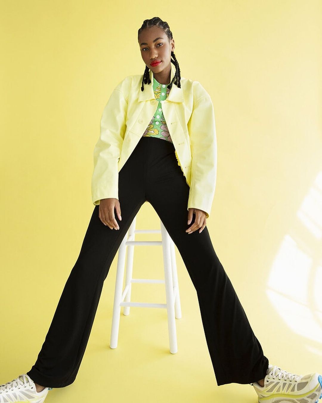 Young woman in a yellow jacket and black pants posing on a white stool against a yellow backdrop in a fashion-style studio portrait.
