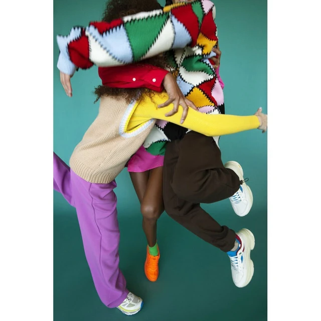 Group of four fashion models embrace, wearing colorful sweaters and sweatpants against a teal backdrop in a studio shot.