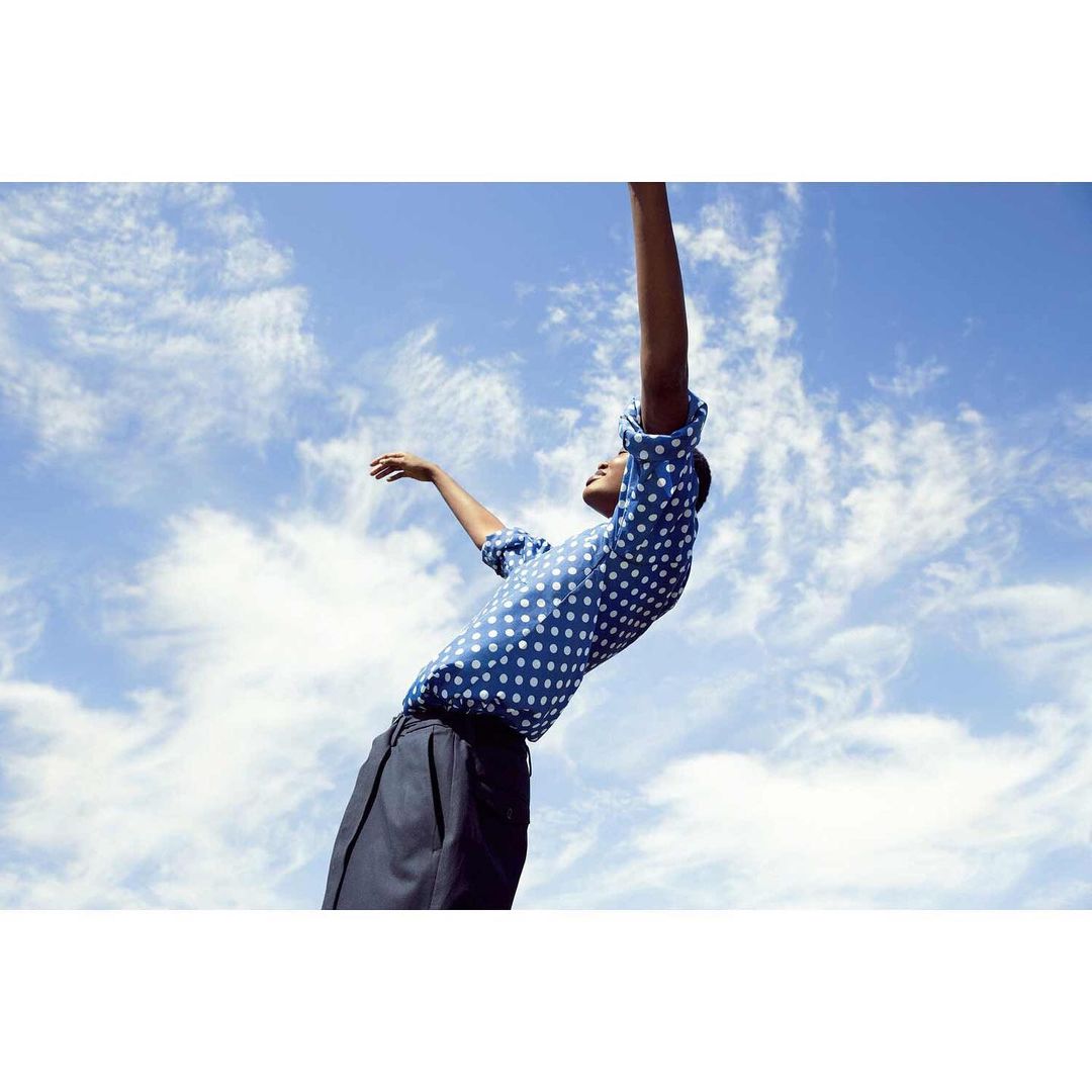Person in polka dot shirt and dark pants with arms raised, against a blue sky with clouds. Fashion shoot, outdoor portrait.