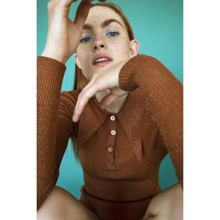 Close-up portrait of a red-haired woman wearing a brown ribbed polo top against a turquoise background.