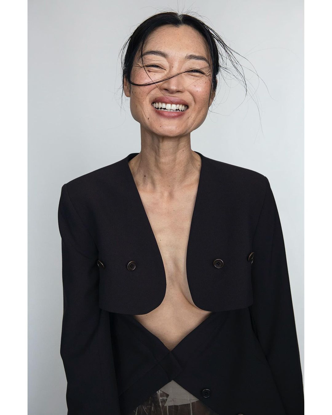 Portrait of an Asian woman smiling, wearing a black blazer against a white backdrop, showcasing editorial fashion and natural beauty.