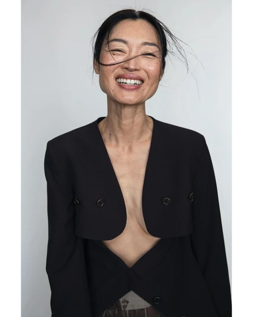 Portrait of an Asian woman smiling, wearing a black blazer against a white backdrop, showcasing editorial fashion and natural beauty.