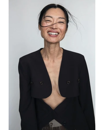 Portrait of an Asian woman smiling, wearing a black blazer against a white backdrop, showcasing editorial fashion and natural beauty.