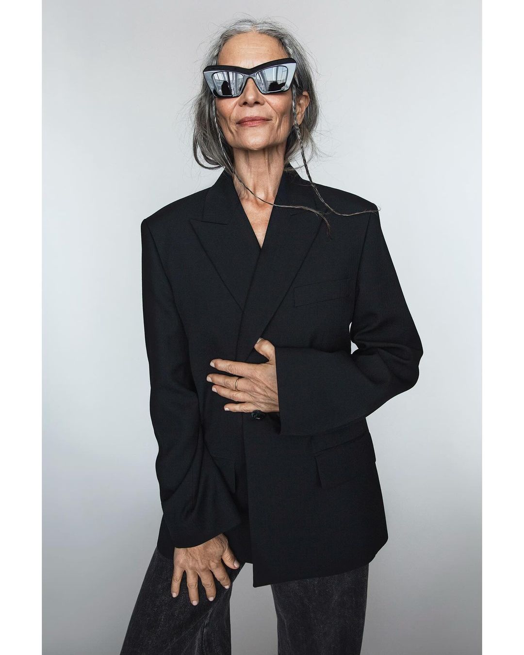 Elegant older woman with gray hair and sunglasses poses in a black blazer against a neutral backdrop, showcasing a sophisticated fashion portrait.