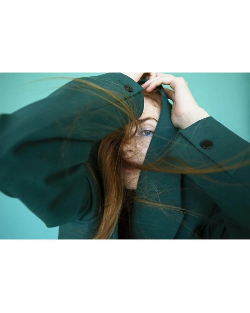 Portrait of a red-haired woman wearing a dark green jacket, holding her hands in her hair against a solid light turquoise background.