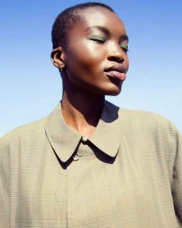 Close-up portrait of a Black woman with short hair wearing a beige plaid collared shirt against a clear blue sky background.
