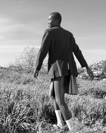 Black and white portrait of a person in a plaid blazer and pleated skirt walking through a tall grass field.