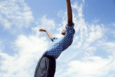 Model in a blue and white polka dot shirt with arms raised against a bright cloudy sky by Brigitte Sire